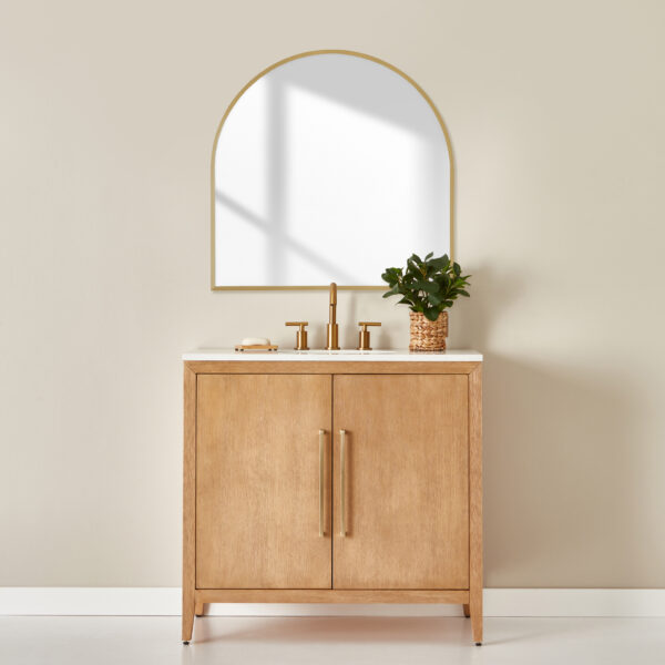 Bathroom vanity with plant on counter. Gold metal accents. Arched shaped mirror hung on wall with bronze frame.