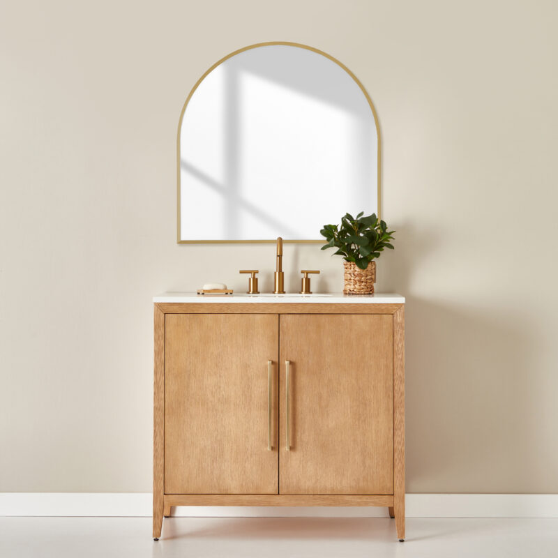 Bathroom vanity with plant on counter. Gold metal accents. Arched shaped mirror hung on wall with bronze frame.
